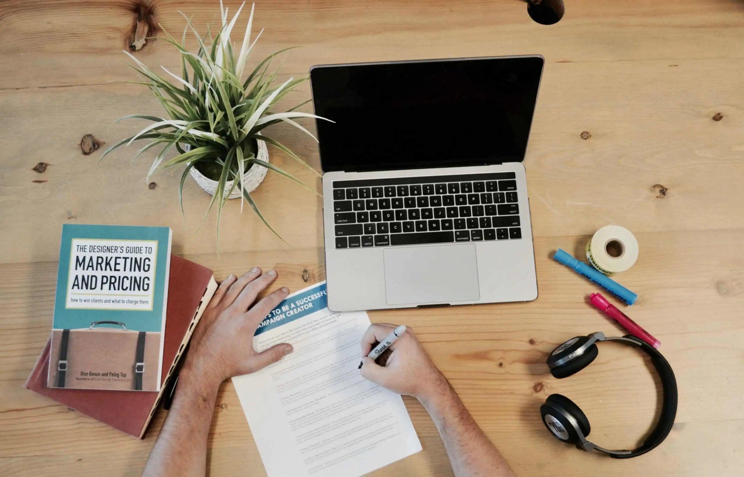 A top-down view of a desk containing a laptop, headphones, a book entitled The Designer's Guide to Marketing, and a man writing on a printed sheet