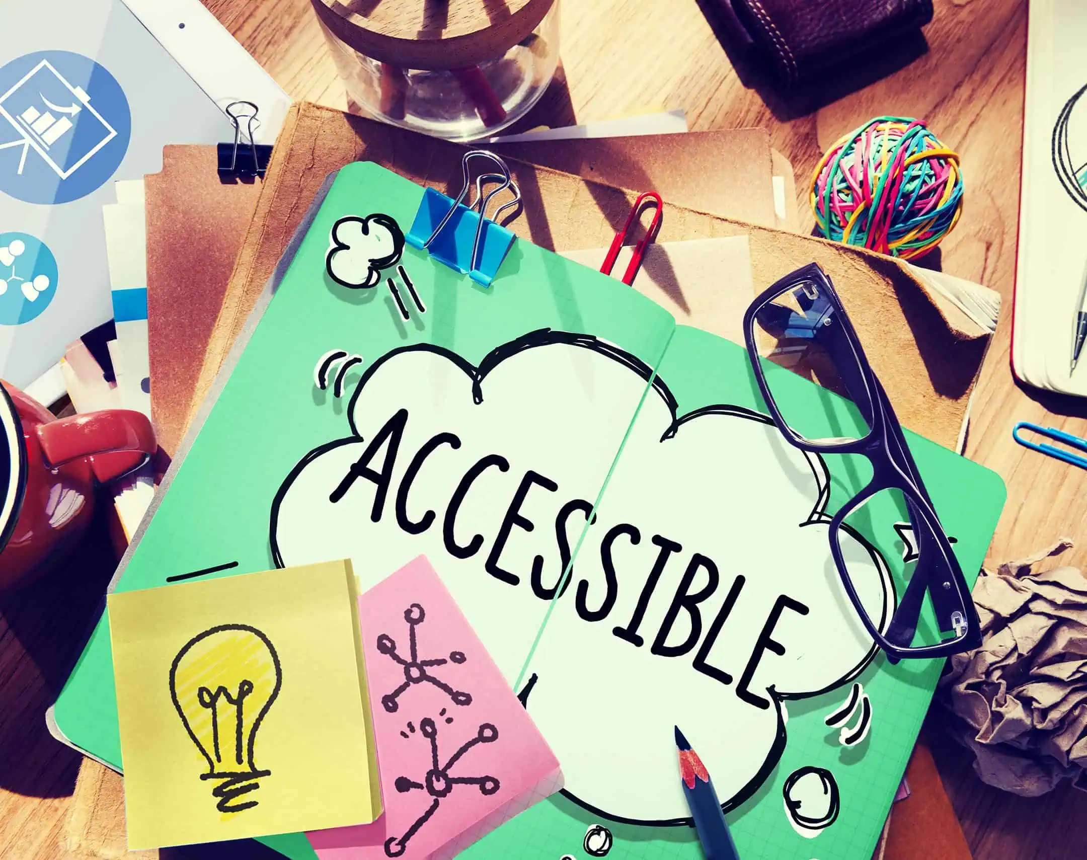 A messy desk including all kinds of items like glasses, elastic bands, paperclips, and a notebook with the word Accessible in a speach bubble