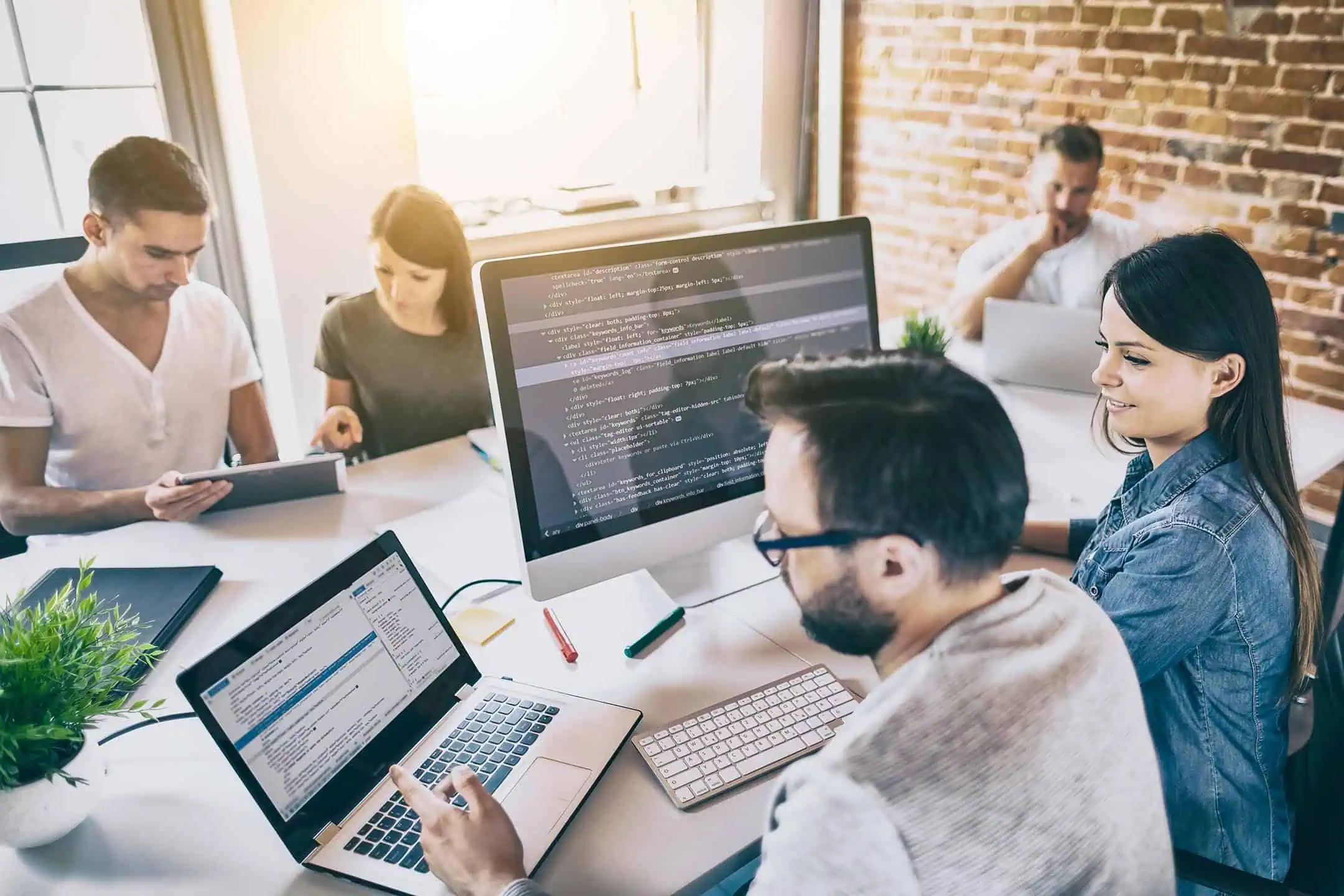 A group of coders sitting around a desk during an office meeting, with code on display on multiple devices atop the table