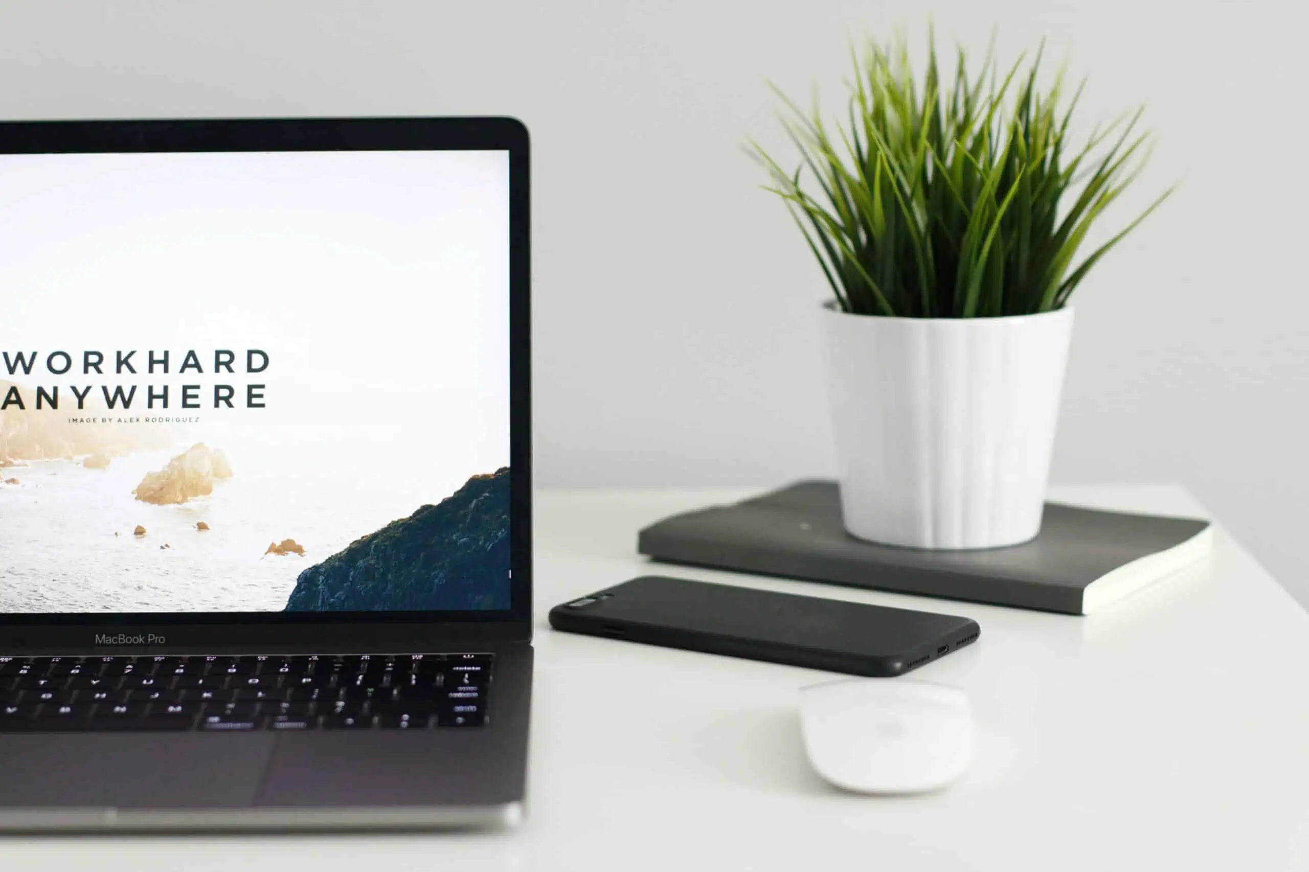 A laptop displaying the text Work Hard Anywhere, beside a mouse, smartphone, and potted plant atop a notebook