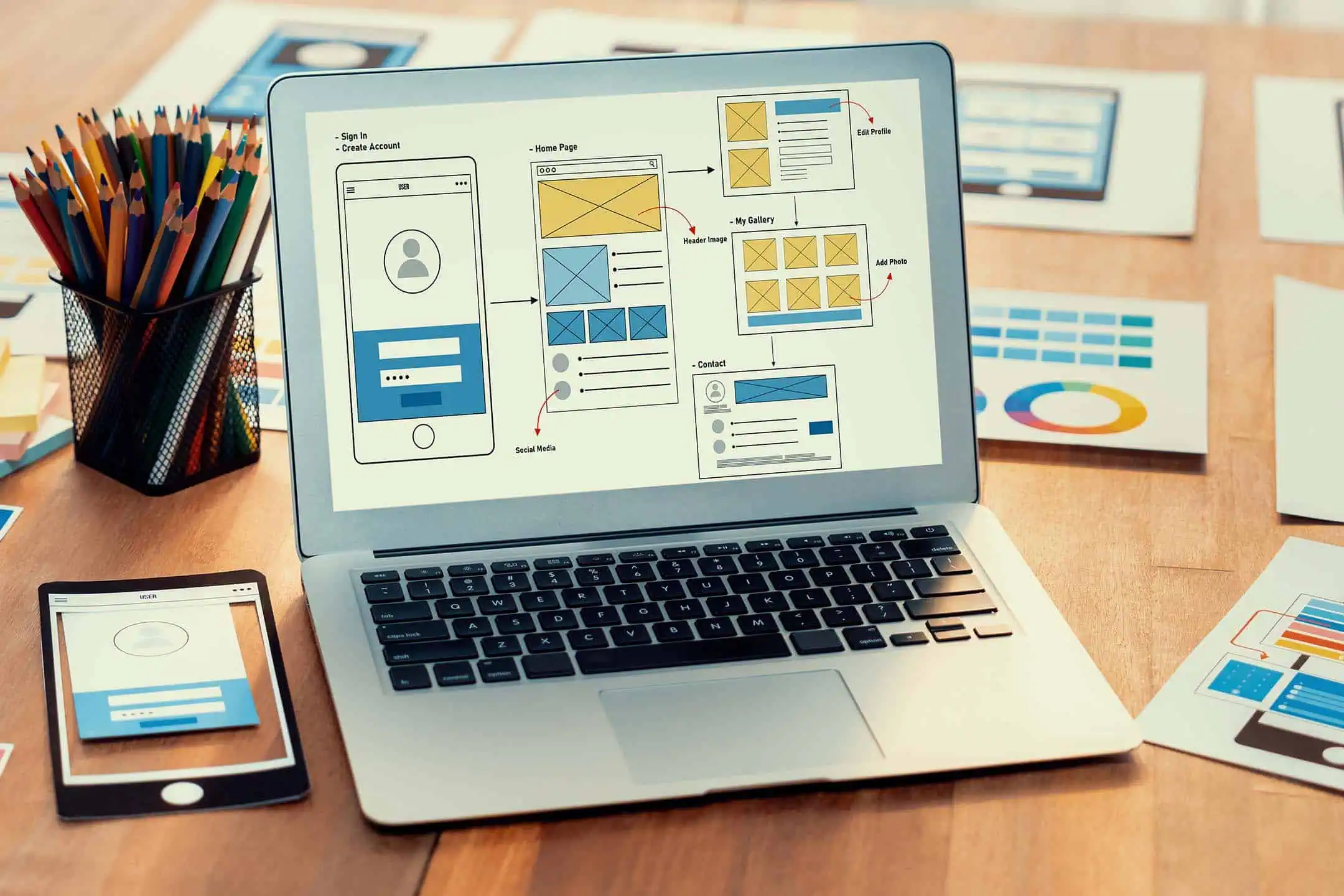 A laptop screen displaying a UI wireframe, surrounded by various printed layouts and concepts